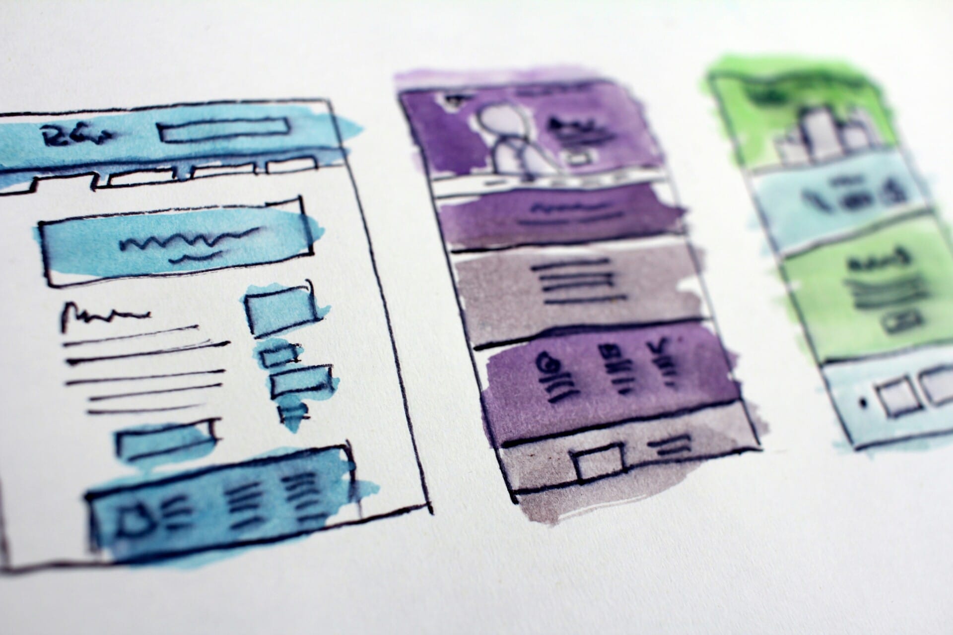 A photo of three sketched website layouts on paper, colored with watercolors in blue, purple, and green. Each layout includes text boxes, image placeholders, and navigation elements. The focus is on design concepts.