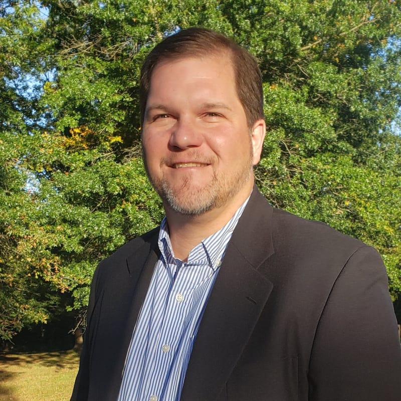 Jeff Tincher from Six10 digital with short brown hair and a beard is smiling outdoors. He is wearing a dark blazer over a light blue striped shirt. The background shows green trees and some patches of sunlight.